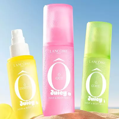 Dos frascos verdes, un perfume rectangular con diseno texturizado y una bruma en formato spray, apoyados sobre la arena de la playa. Una toalla de playa doblada a rayas rosas descansa junto a los frascos. De fondo, el mar y un cielo azul despejado.