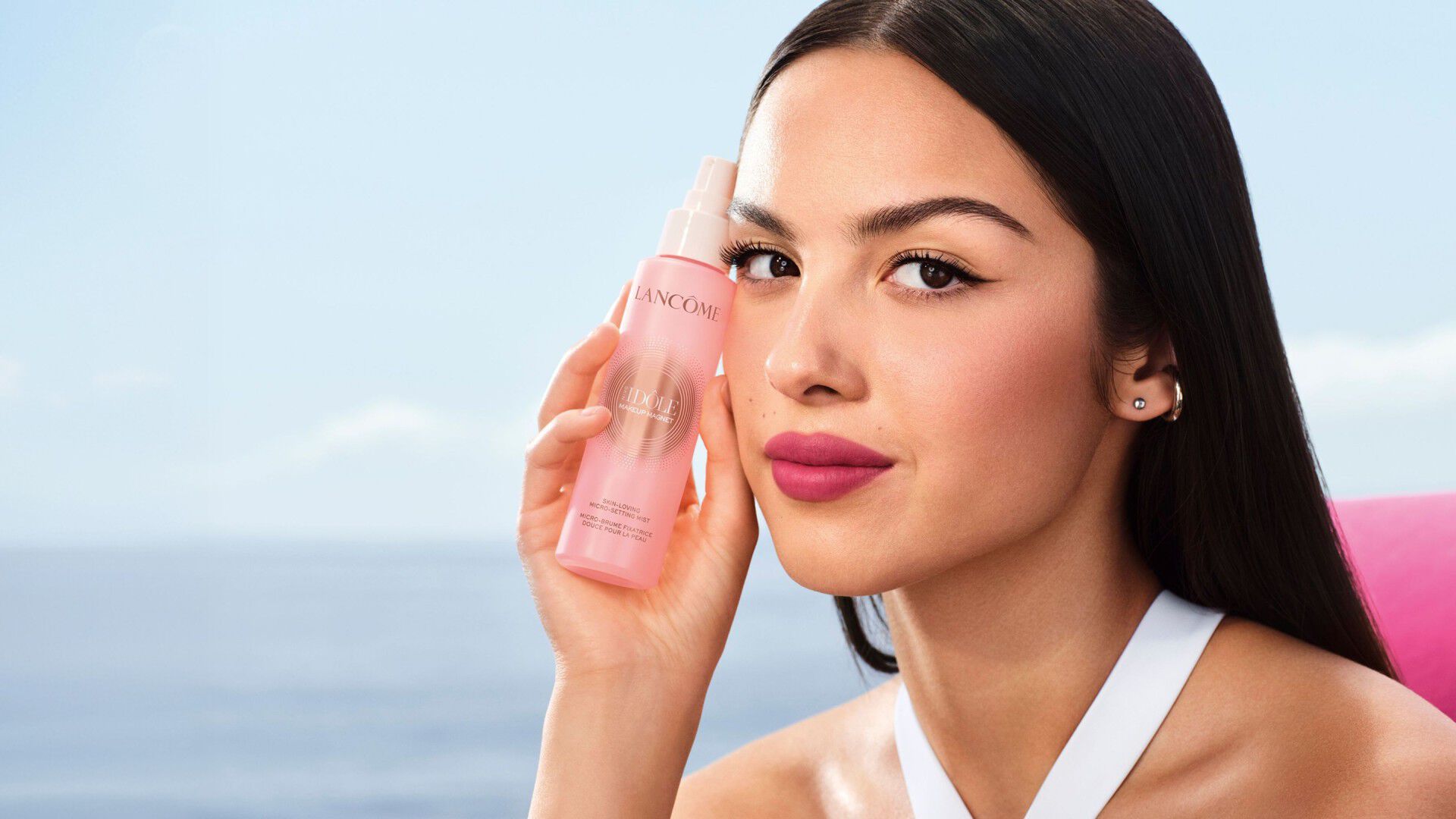 Modelo sosteniendo la Micro Bruma Fijadora Skin Idôle Makeup Magnet de Lancôme junto al mar.
